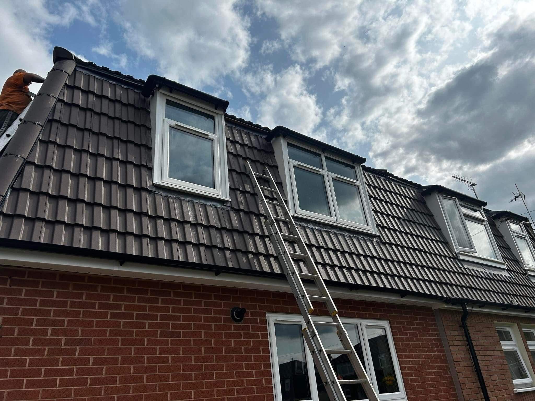 A person works near the edge of a tiled roof on a two-story brick house, with a ladder leaning against the roof and clouds visible in the sky.