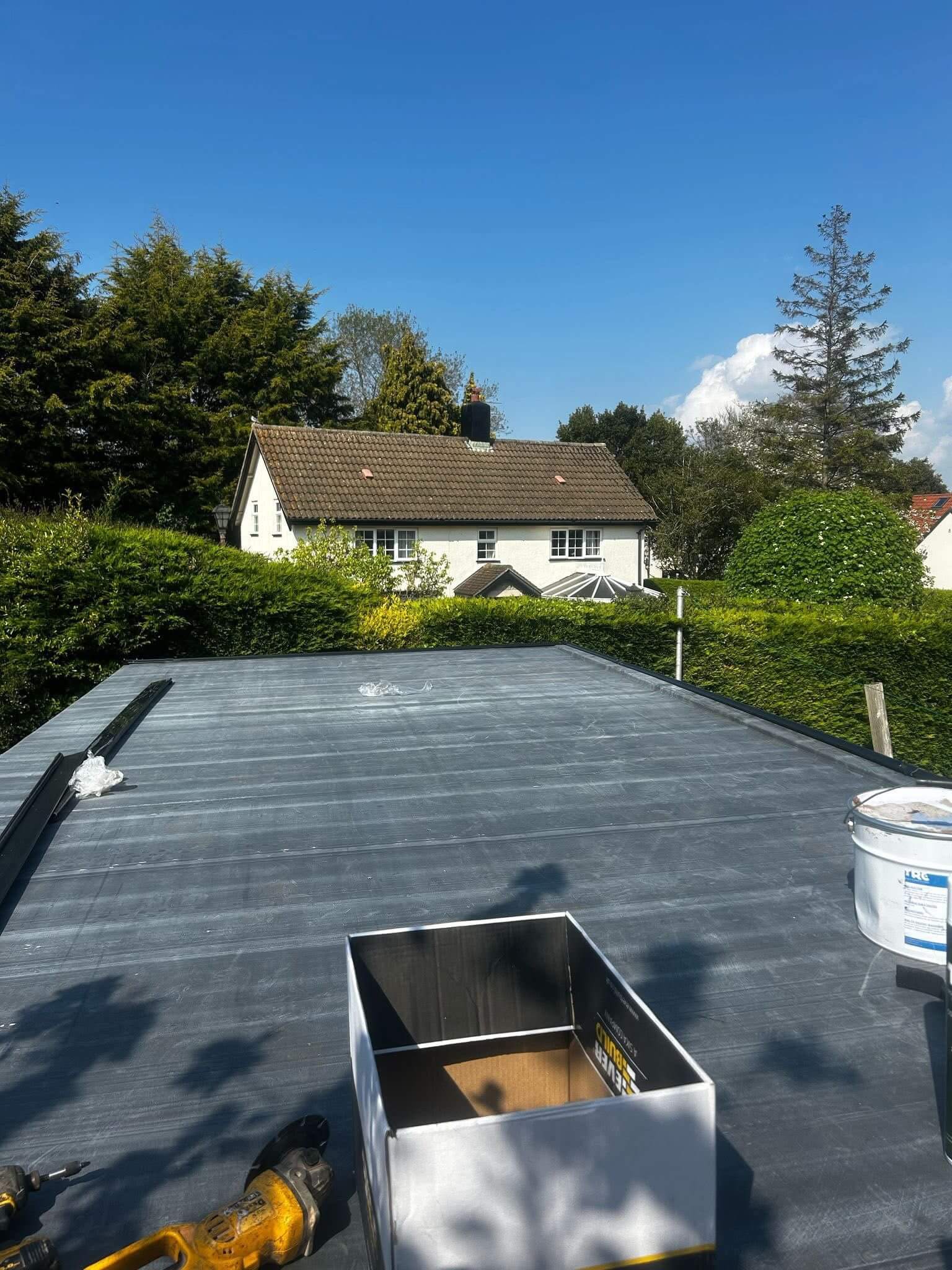 Flat roof under construction with tools, a box, and a paint bucket in the foreground; a house and trees are visible in the background on a sunny day.