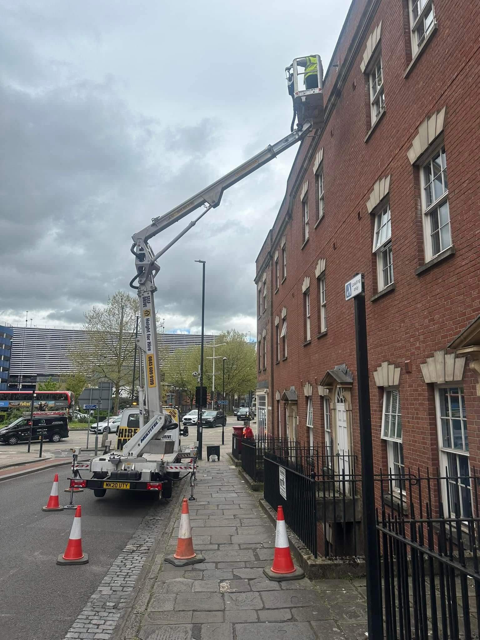 A worker in a lift is elevated to the upper floor of a brick building on a city street, with safety cones placed around the lift truck.