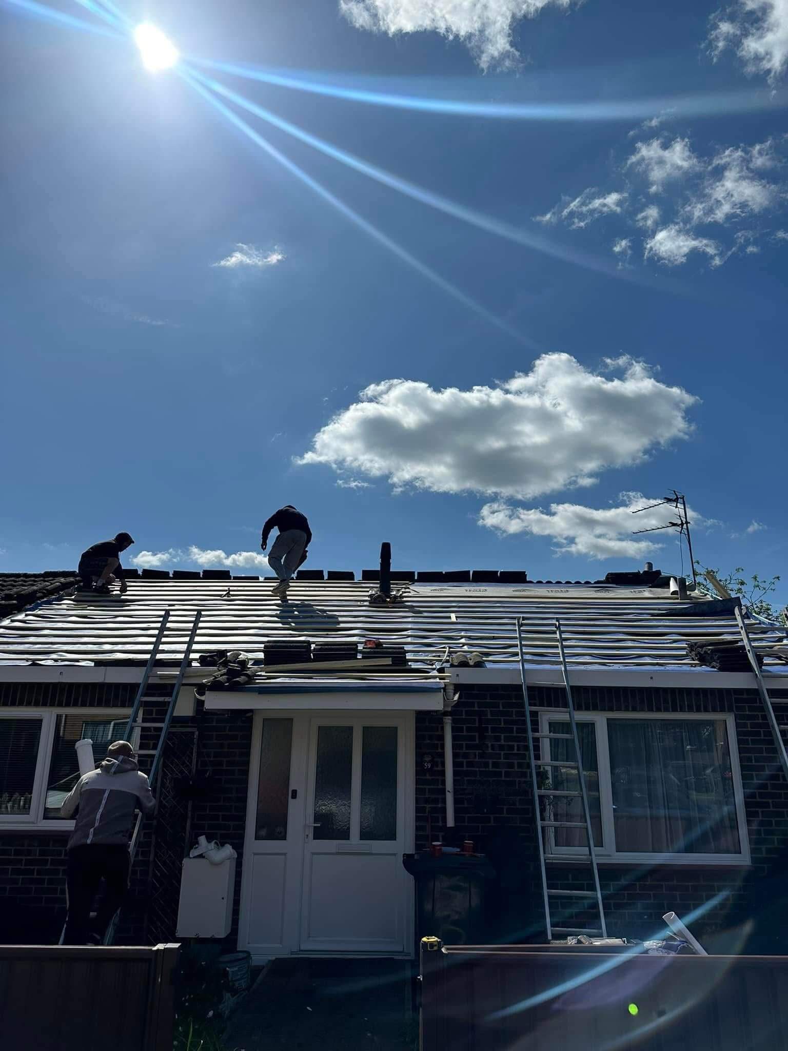 Three people work on the roof of a house under a bright blue sky with scattered clouds and sunlight streaming down.
