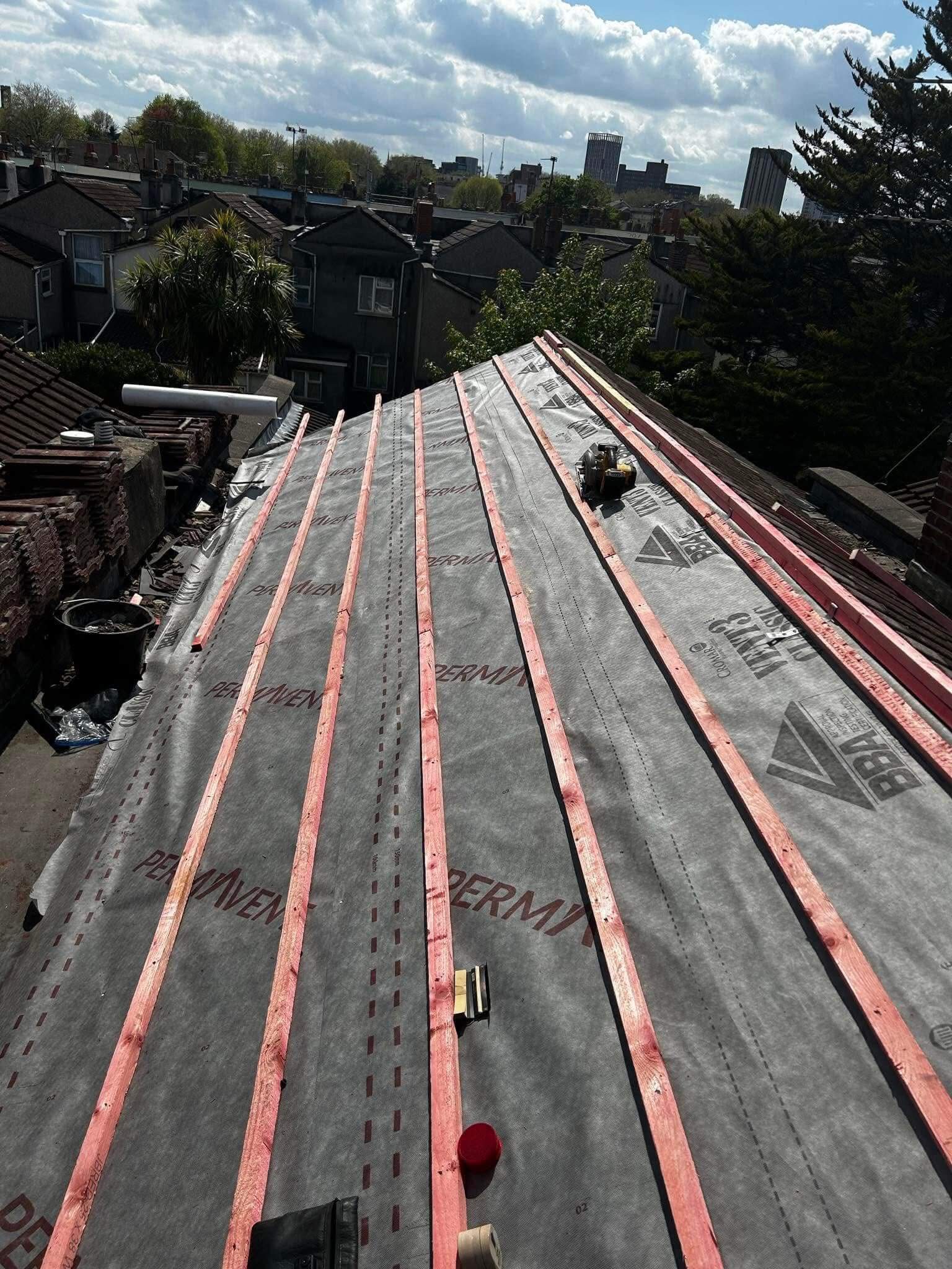 A sloped roof under construction with waterproof membrane and red wooden battens installed; tools and materials are scattered on the surface. Houses and trees are visible in the background.