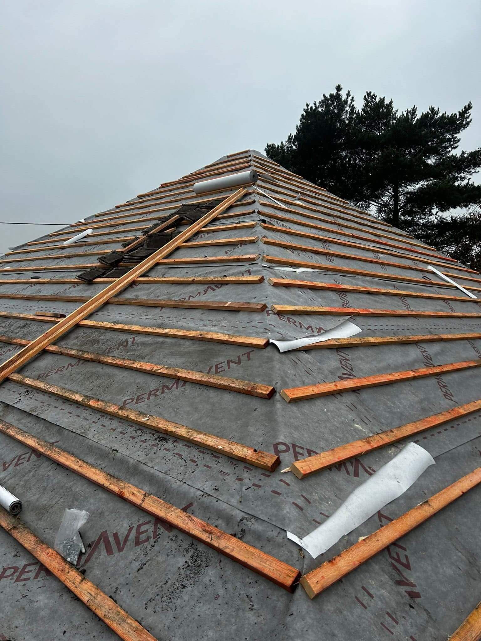 A sloped roof under construction with wooden battens fixed over a waterproof membrane, set against a cloudy sky and trees in the background.