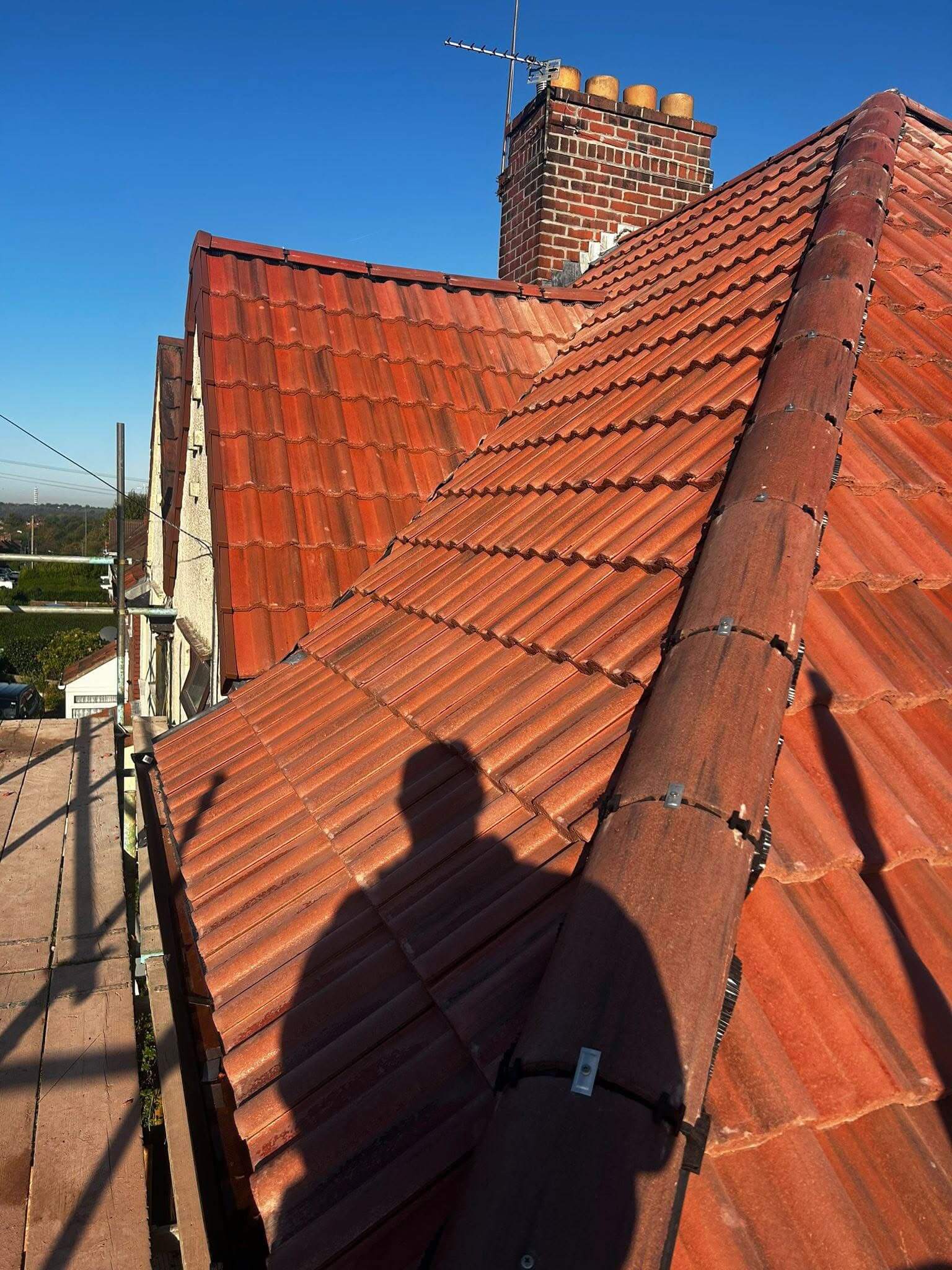 Red tiled roof with a chimney in the background and a person’s shadow cast on the tiles under clear blue sky.