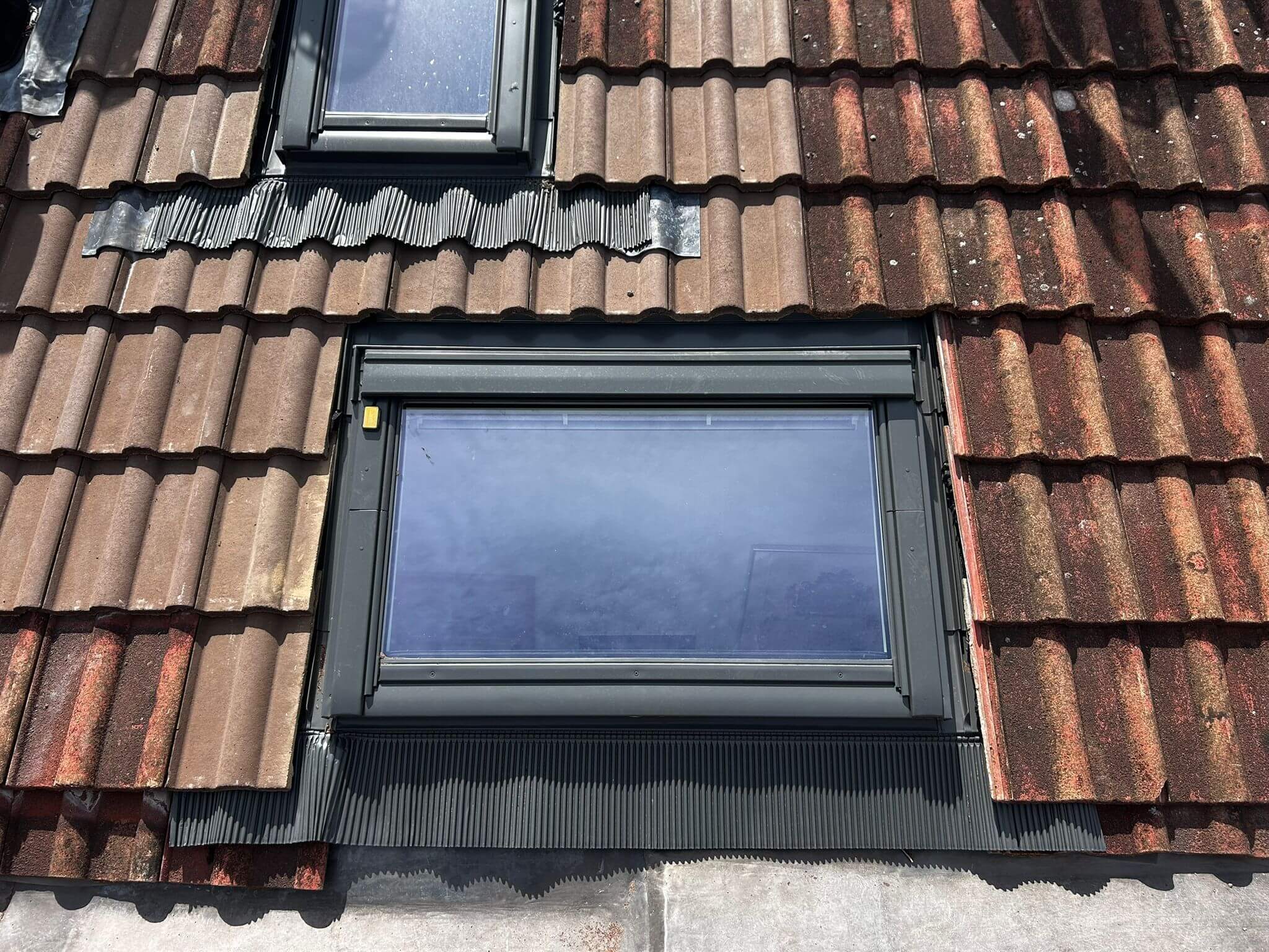 A close-up view of a house roof with brown tiles featuring two rectangular skylight windows framed in black.