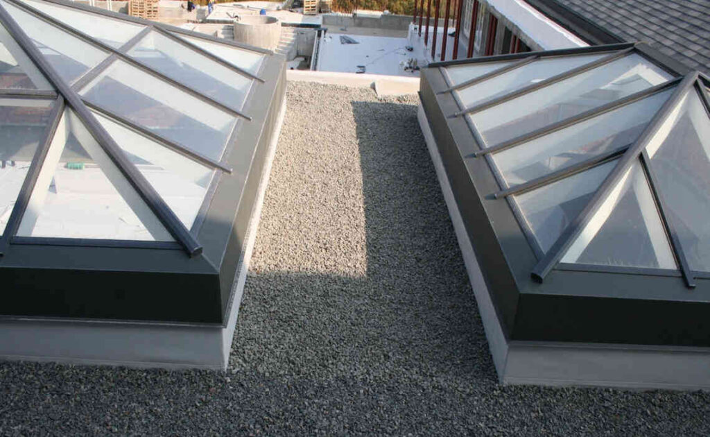Two large glass skylights with metal frames are installed on a gravel-covered flat rooftop, adjacent to areas where you'll need to repair fascia and soffit for optimal durability.