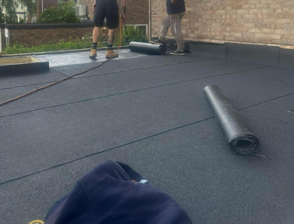 Two workers are installing roofing felt on a flat rooftop; rolls of material and tools for roof and repair fascia and soffit work are visible on the surface.