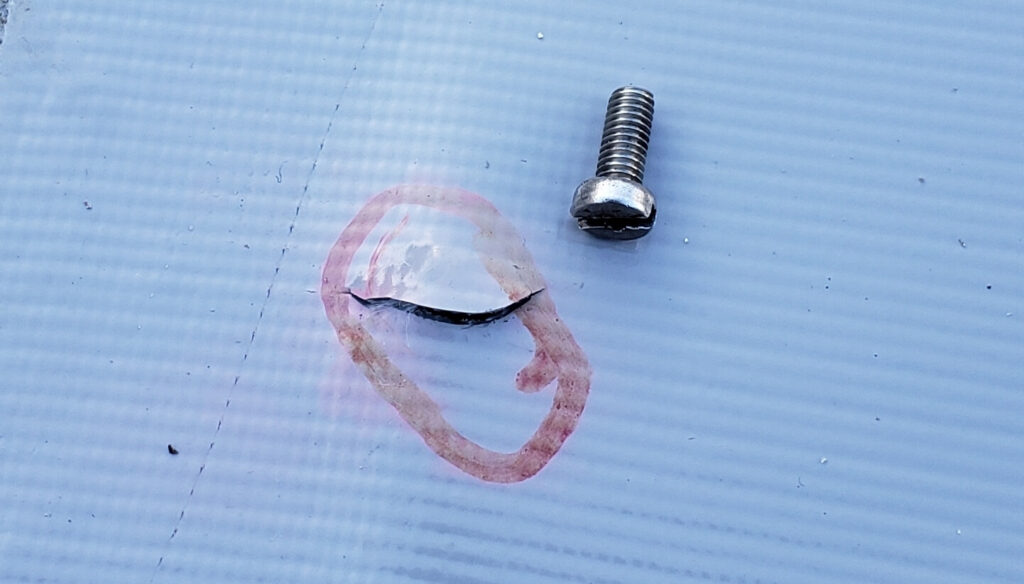 A metal screw sits next to a cracked plastic piece with a reddish circular stain on a textured white surface, possibly remnants from repair fascia and soffit work.